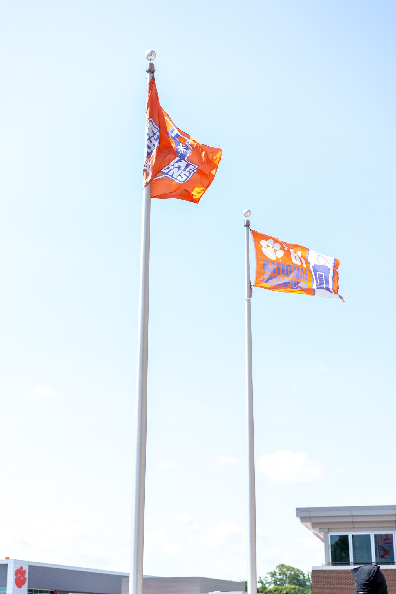Two orange Clemson Tigers flags flying on tall flagpoles against a clear blue sky near a brick building.