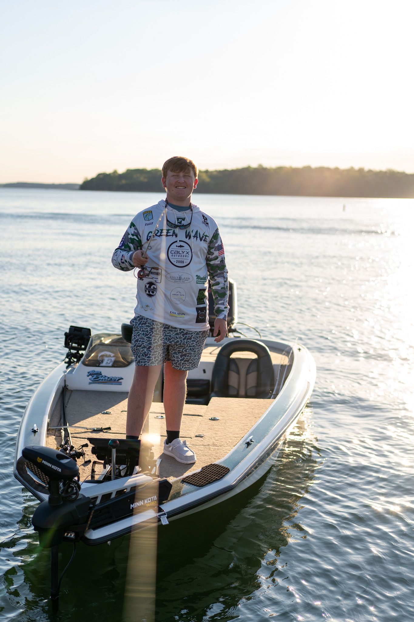 Young angler stands on a fishing boat at sunset wearing a Green Wave jersey and camo shorts on calm lake waters.