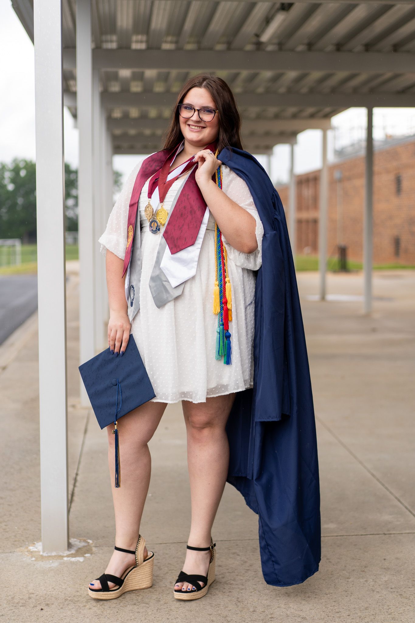 A smiling young woman in graduation regalia with navy gown, mortarboard, and colorful honor cords poses outdoors.