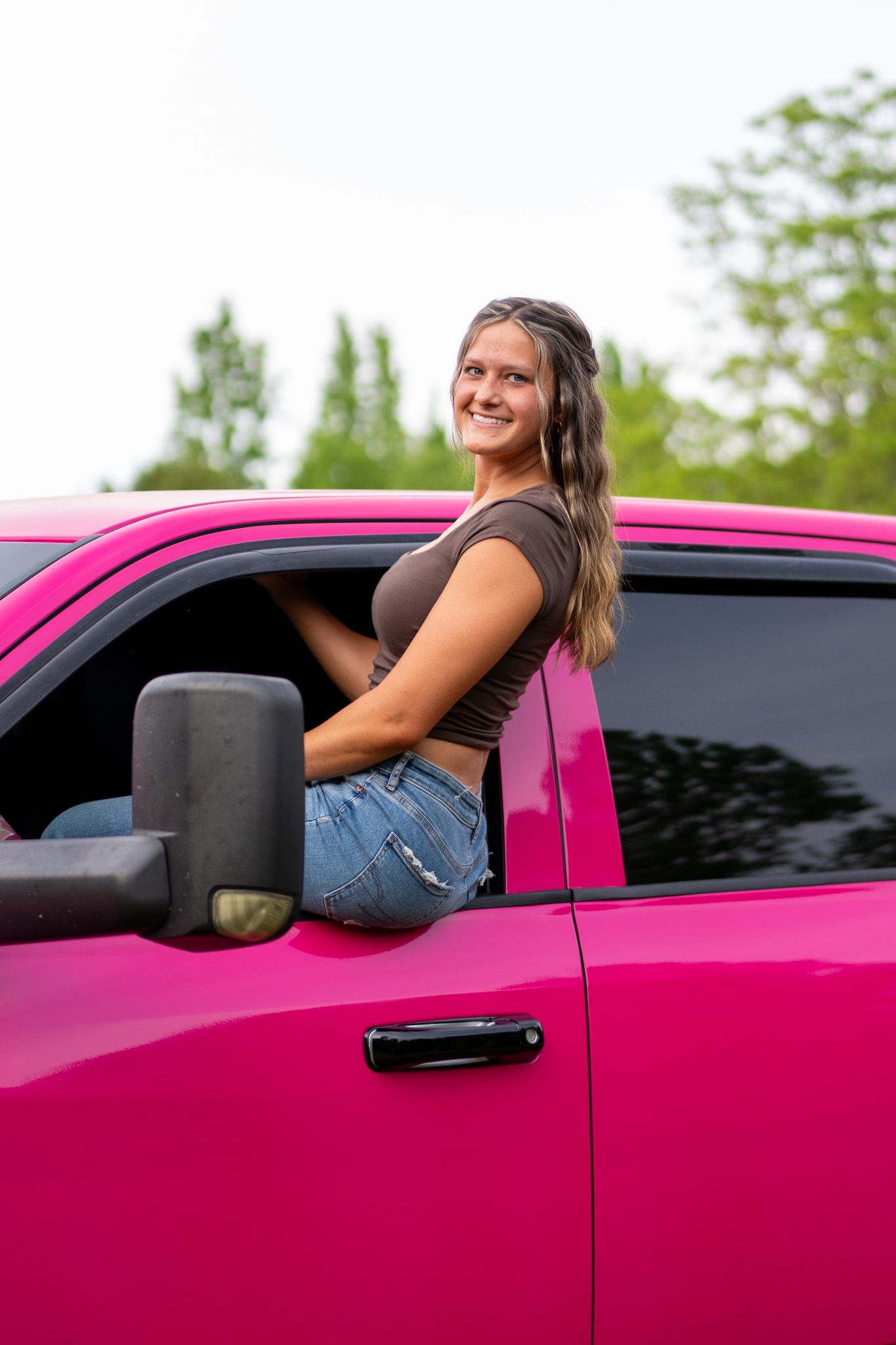 Young woman in brown top and denim shorts smiling beside a vibrant pink pickup truck outdoors.