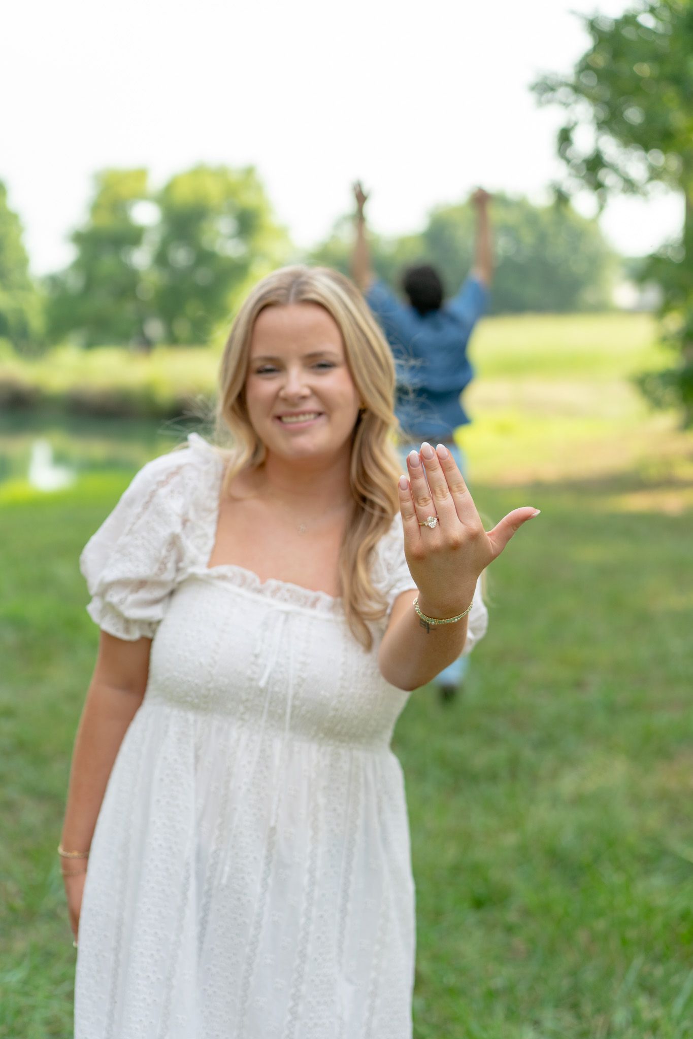 Smiling blonde woman in white dress showing engagement ring while man celebrates joyfully behind her.