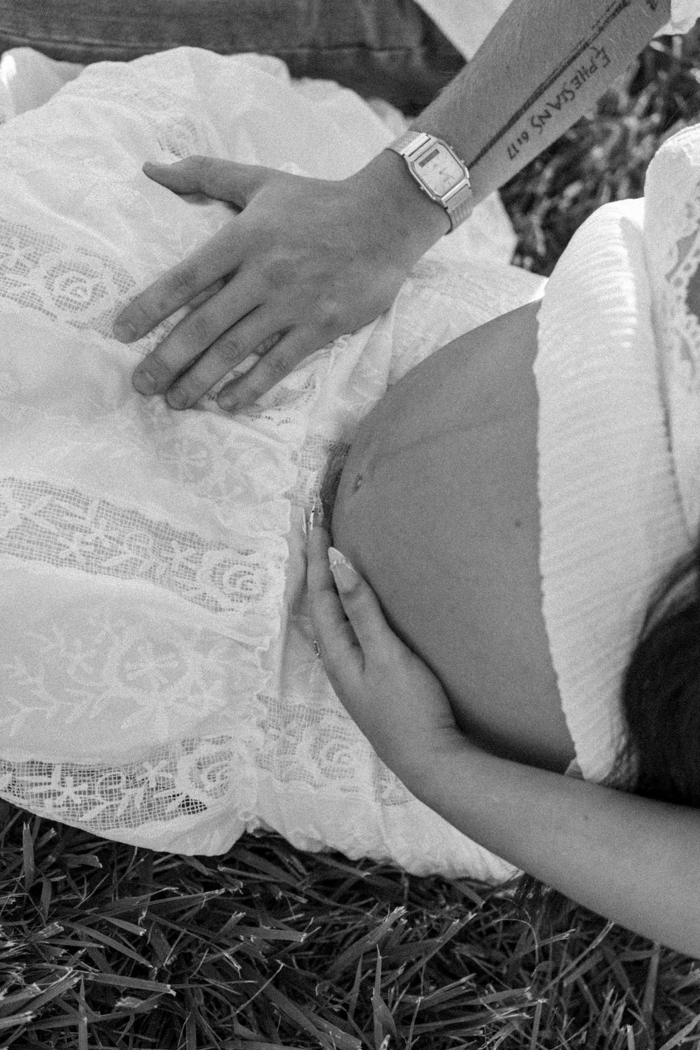Black and white photo of pregnant woman in white lace dress with partner's hand resting on her baby bump.