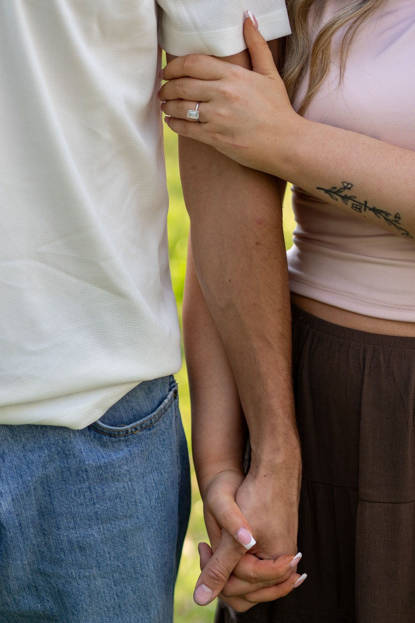 Close-up of couple holding hands with engagement ring and floral tattoo visible outdoors.