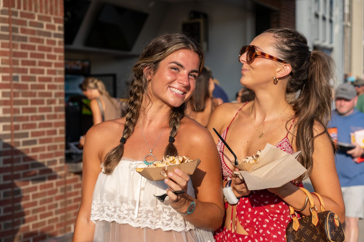 Two smiling women holding food containers outdoors at a summer festival event.