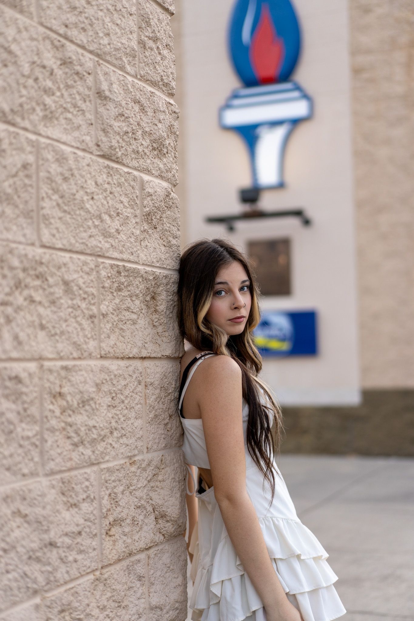 Young woman in white ruffled dress leaning against brick wall outdoors near a torch sign.