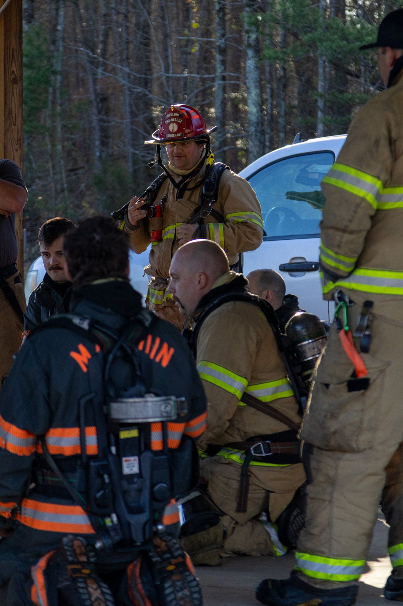 Firefighters in turnout gear with SCBA tanks gather outdoors near trees during an emergency response or training exercise.
