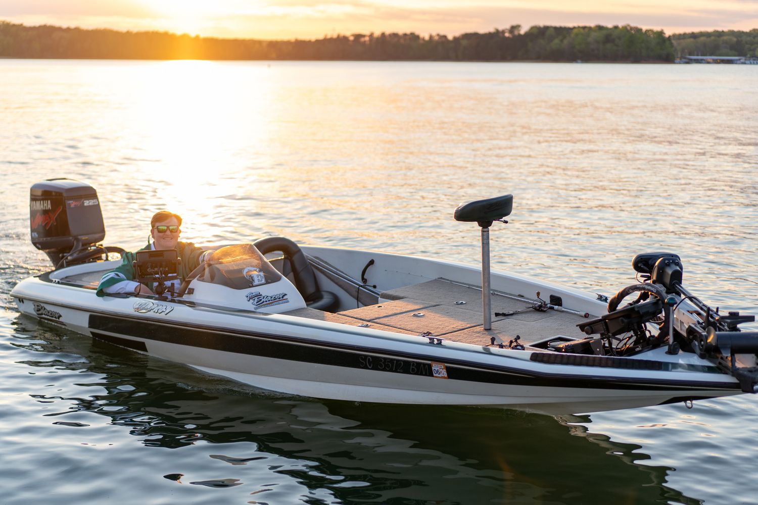 A sleek white bass fishing boat glides across calm water at golden sunset with an angler seated at the helm.