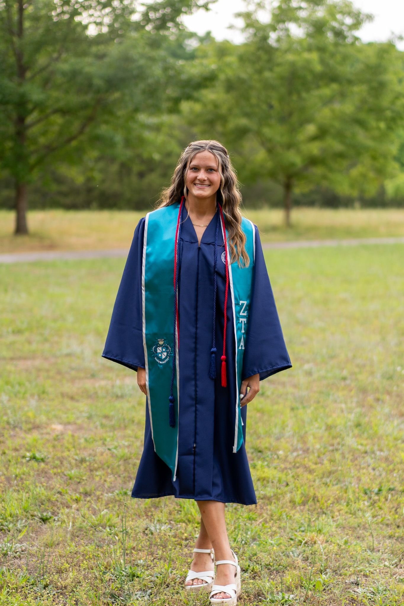 Smiling graduate in navy blue cap and gown with teal stole standing outdoors in grassy park setting.