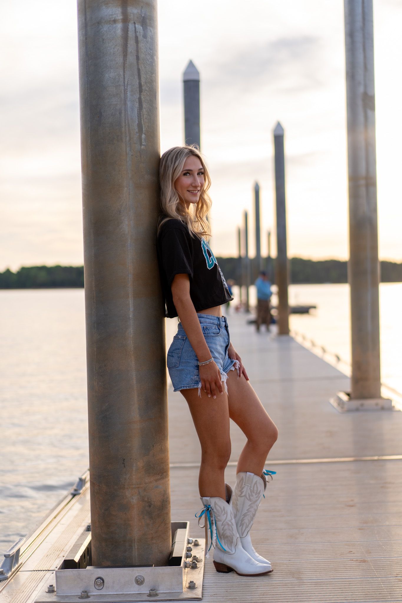 Smiling blonde woman in denim shorts and white cowboy boots leaning against a pier post at sunset.