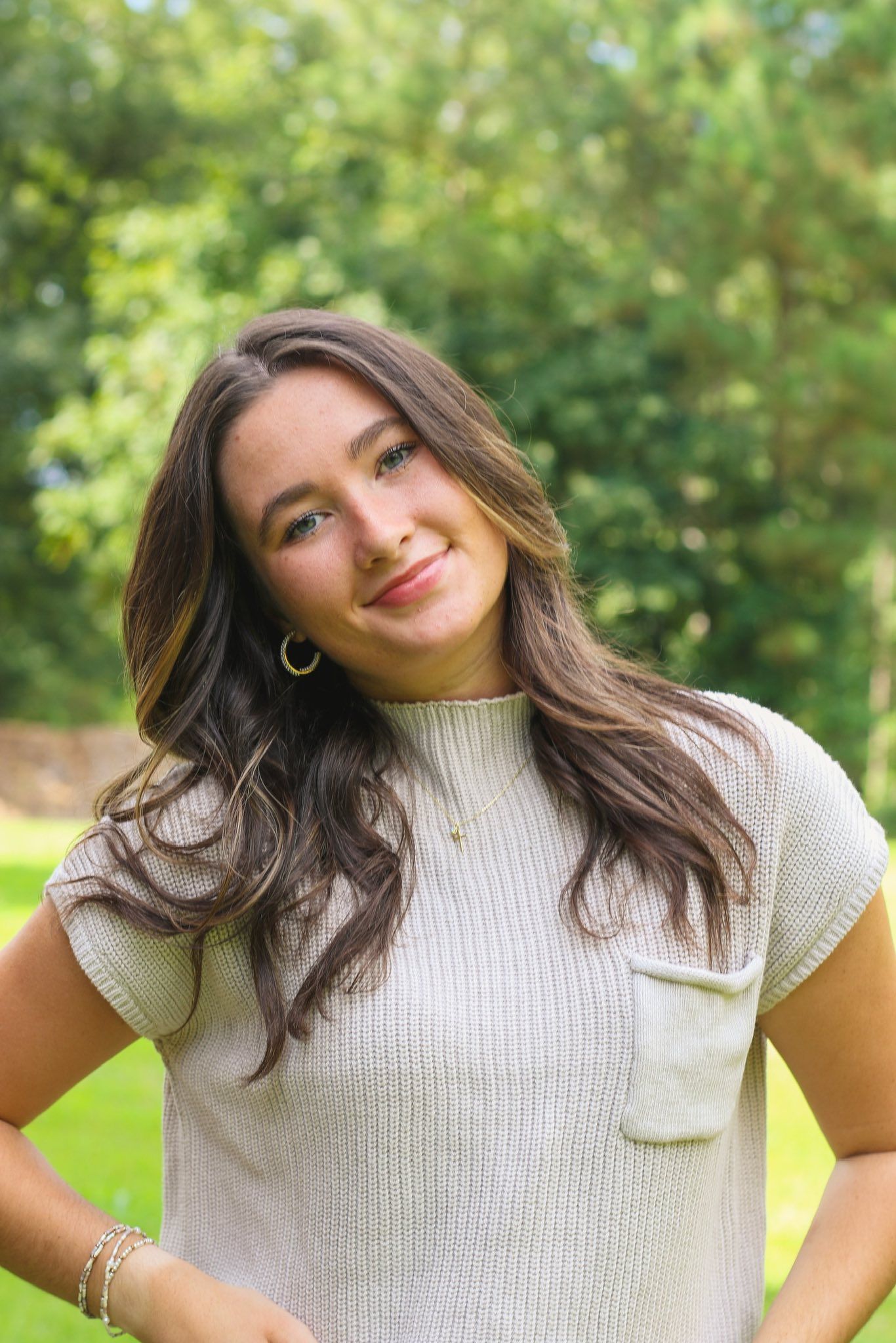 Smiling brunette woman in cream ribbed top posing outdoors in green park setting.