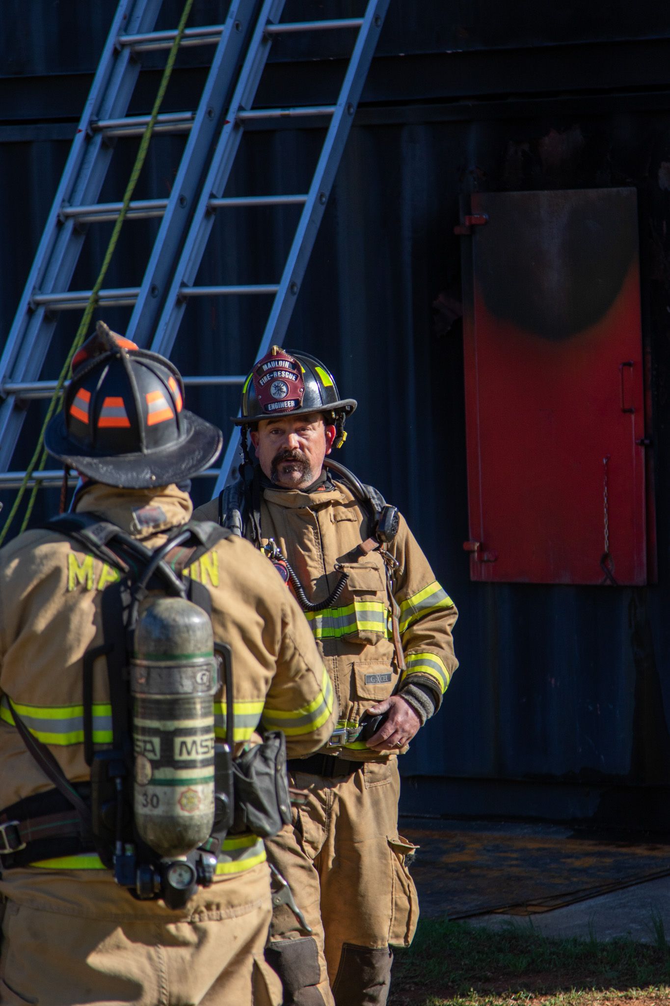 Two firefighters in full protective gear discuss tactics near a training building with a ladder and red door.