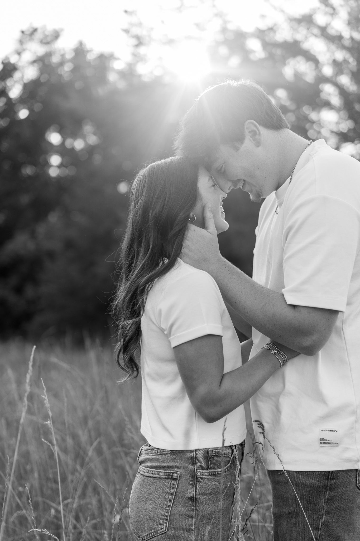 Black and white romantic photo of a young couple nearly kissing in a sun-drenched grassy field.