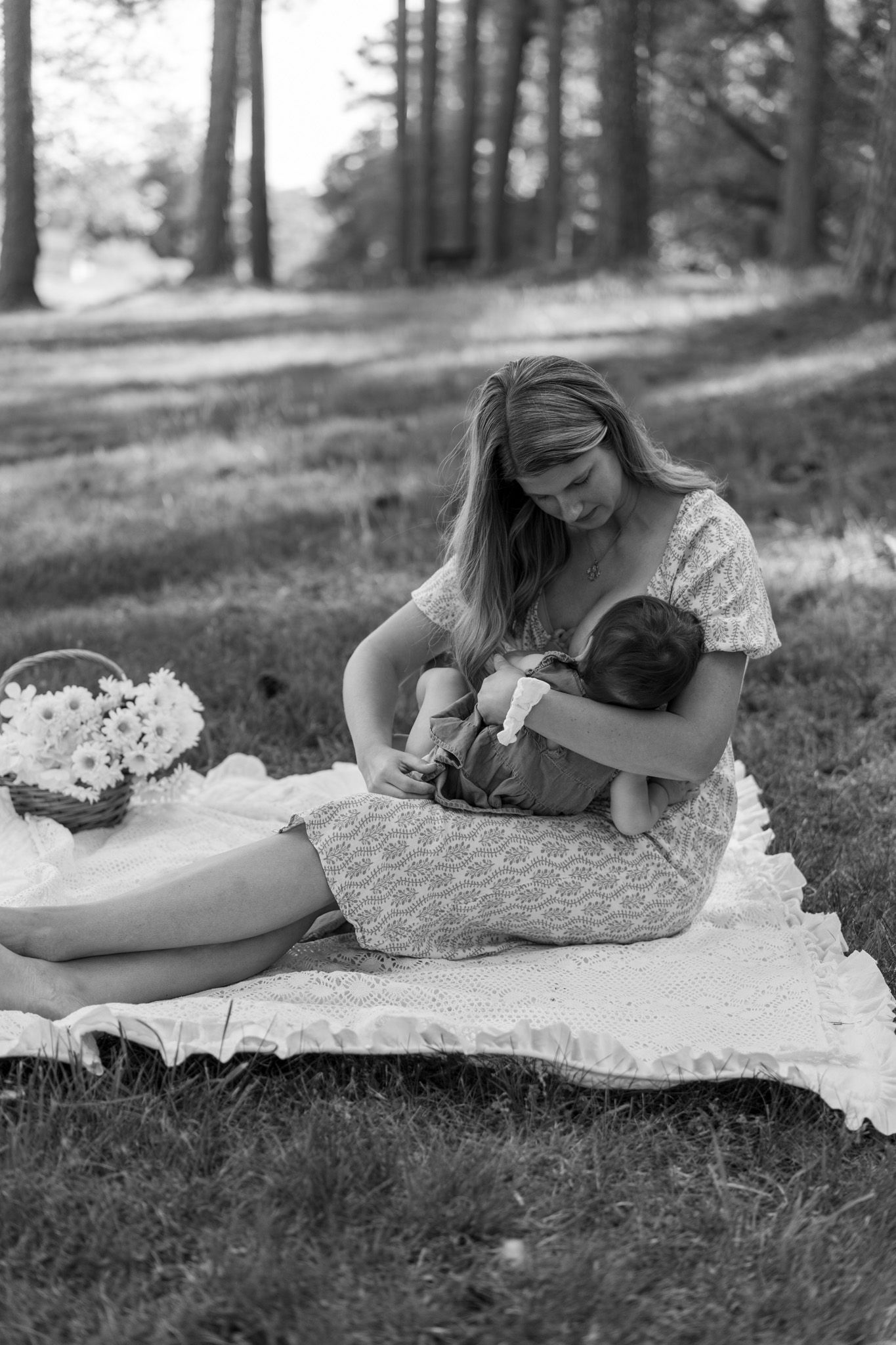 Black and white photo of blonde mother cradling sleeping newborn on blanket in peaceful wooded park.