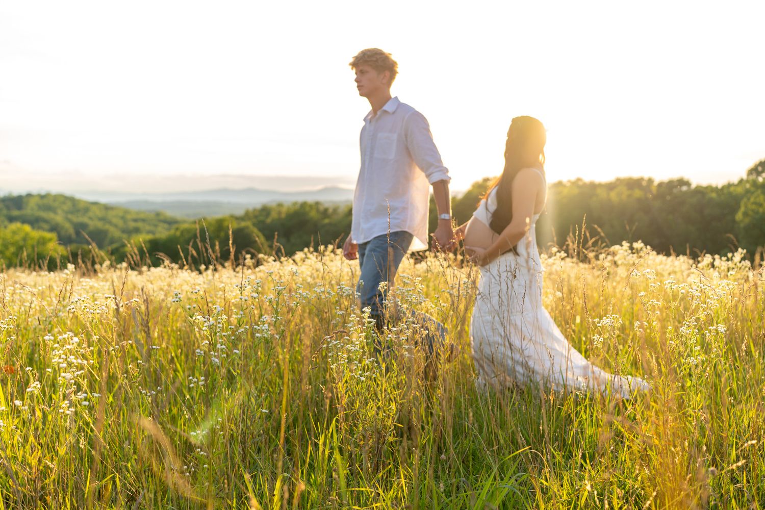 Expecting couple holding hands while walking through golden wildflower field during stunning sunset.