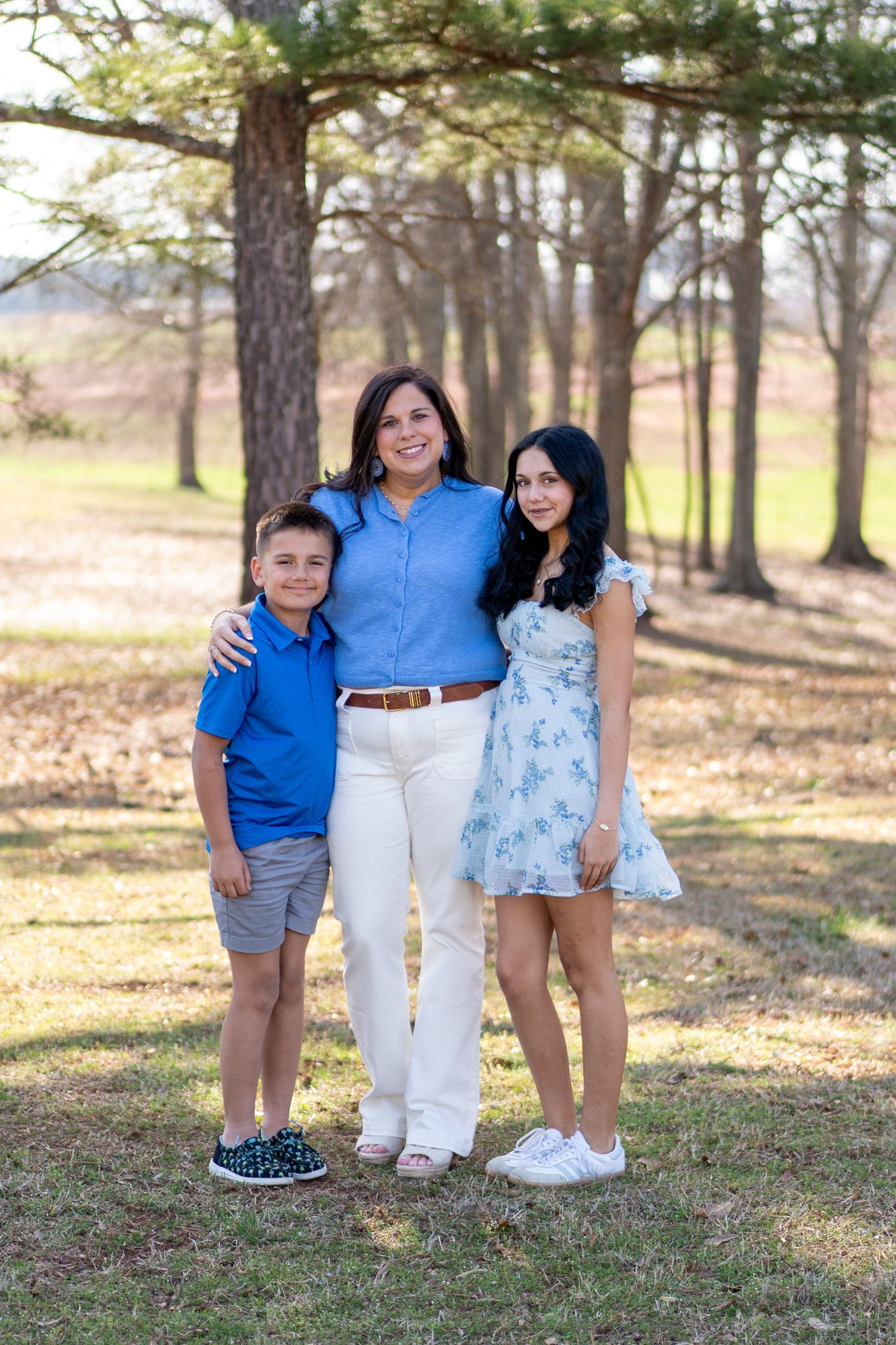 A woman in a blue shirt stands between her two children, smiling for a family portrait in a wooded park setting.