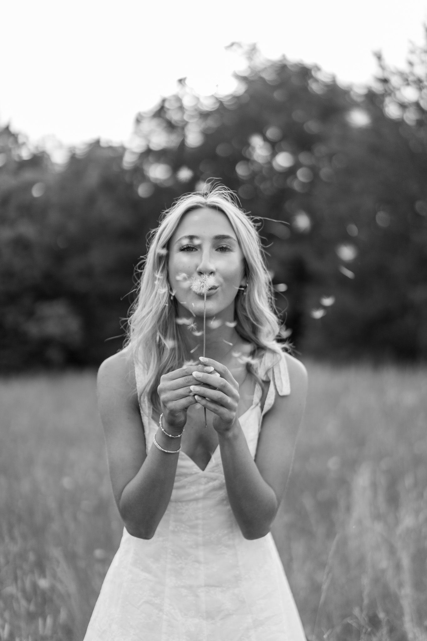 Young blonde woman blowing dandelion seeds in a field, captured in black and white.