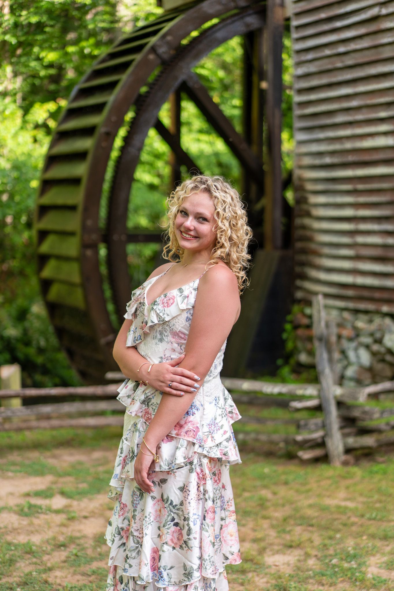 Young blonde woman in floral dress smiling beside rustic wooden water wheel mill outdoors.