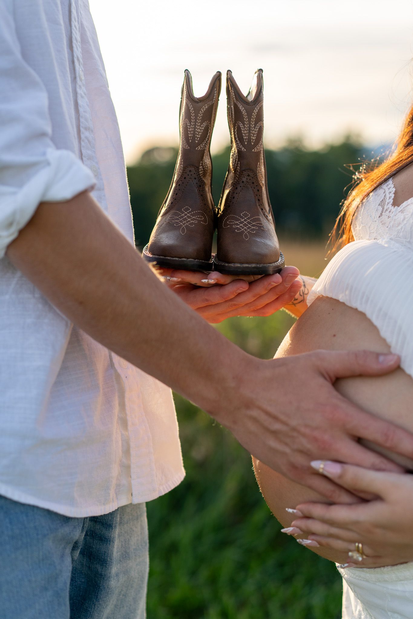 Couple holding tiny cowboy boots near pregnant belly in a field at sunset, maternity photo session.