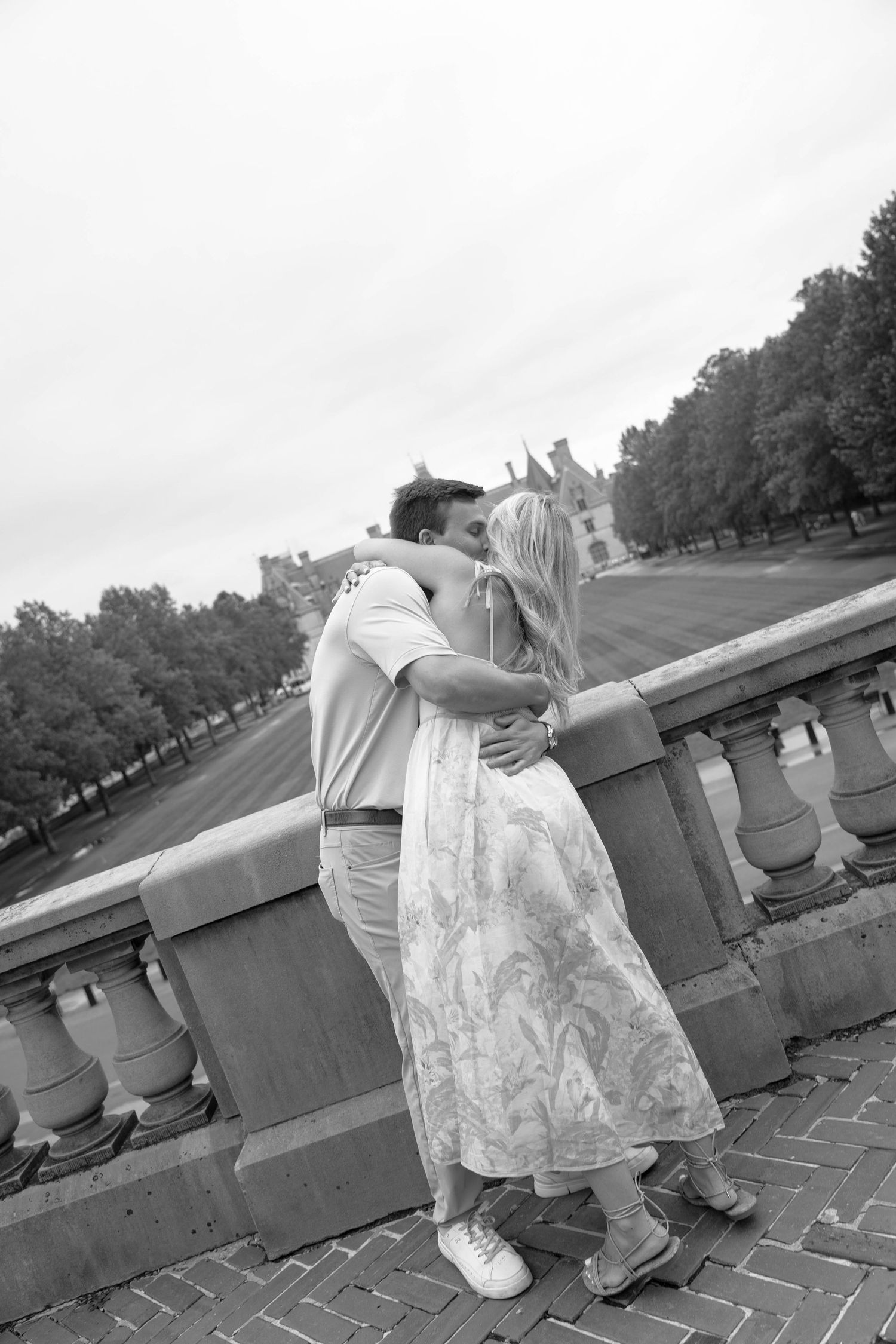 Black and white photo of couple kissing by stone balustrade with historic estate in background.
