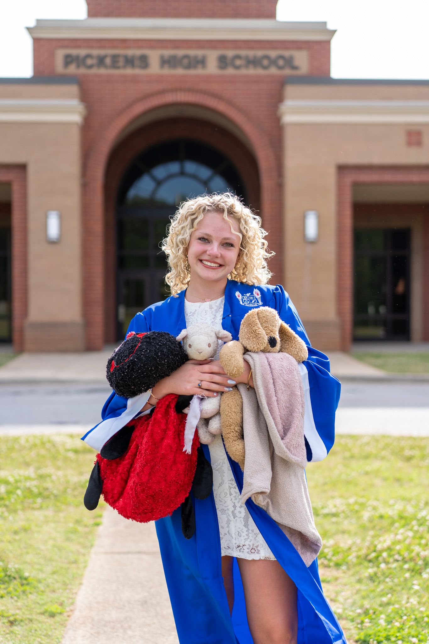 Smiling high school graduate in blue gown holding stuffed animals outside Pickens High School.