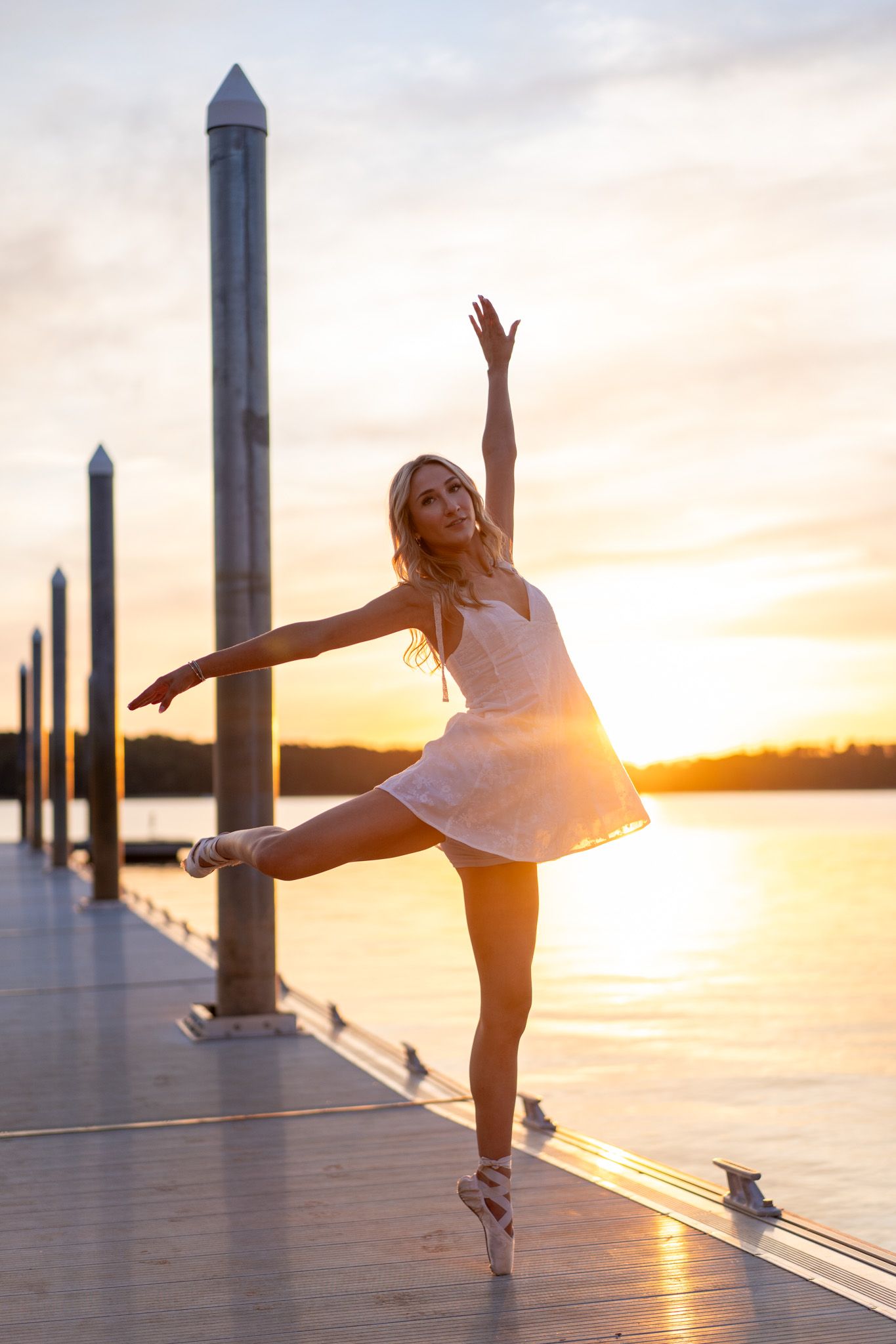 Graceful ballerina in white dress performing on a pier during a stunning golden hour sunset.