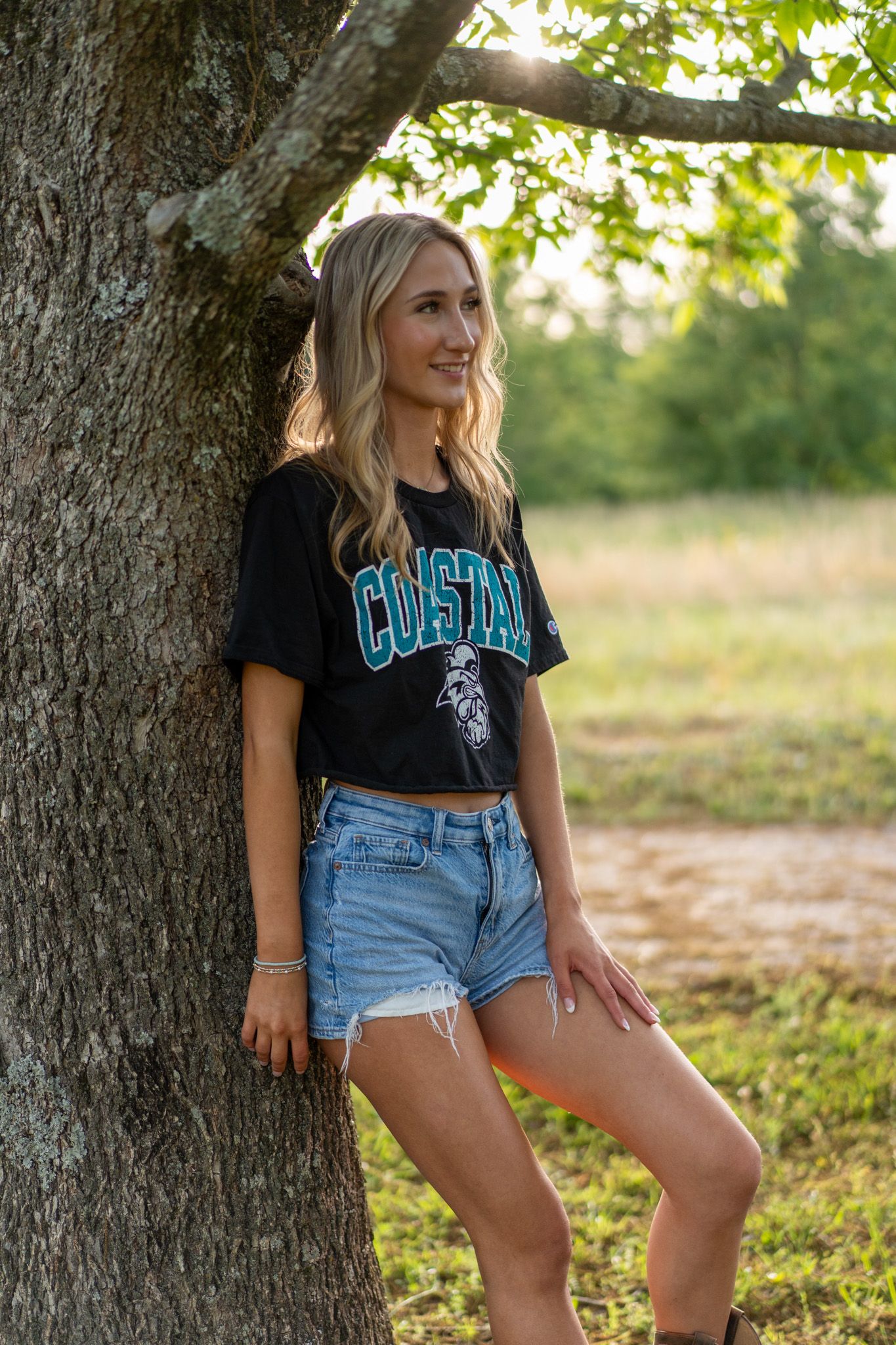 Young blonde woman in a black Coastal crop tee and denim cutoff shorts leaning against a tree outdoors.
