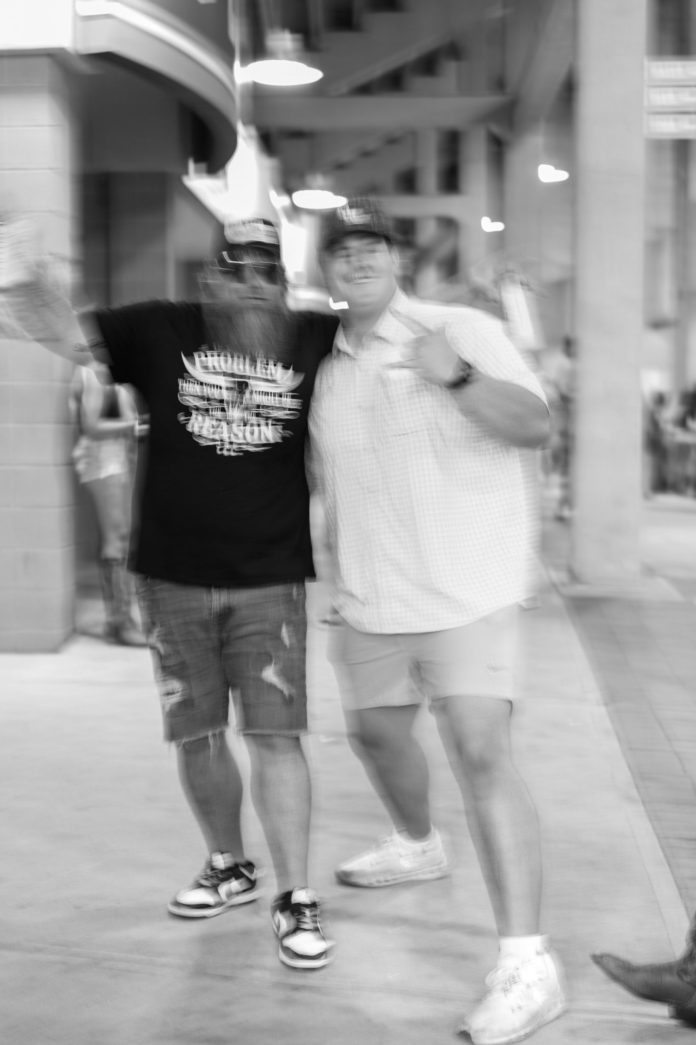 Two men pose together smiling on a busy street, one in a black graphic tee and one in a white shirt, in black and white photo.