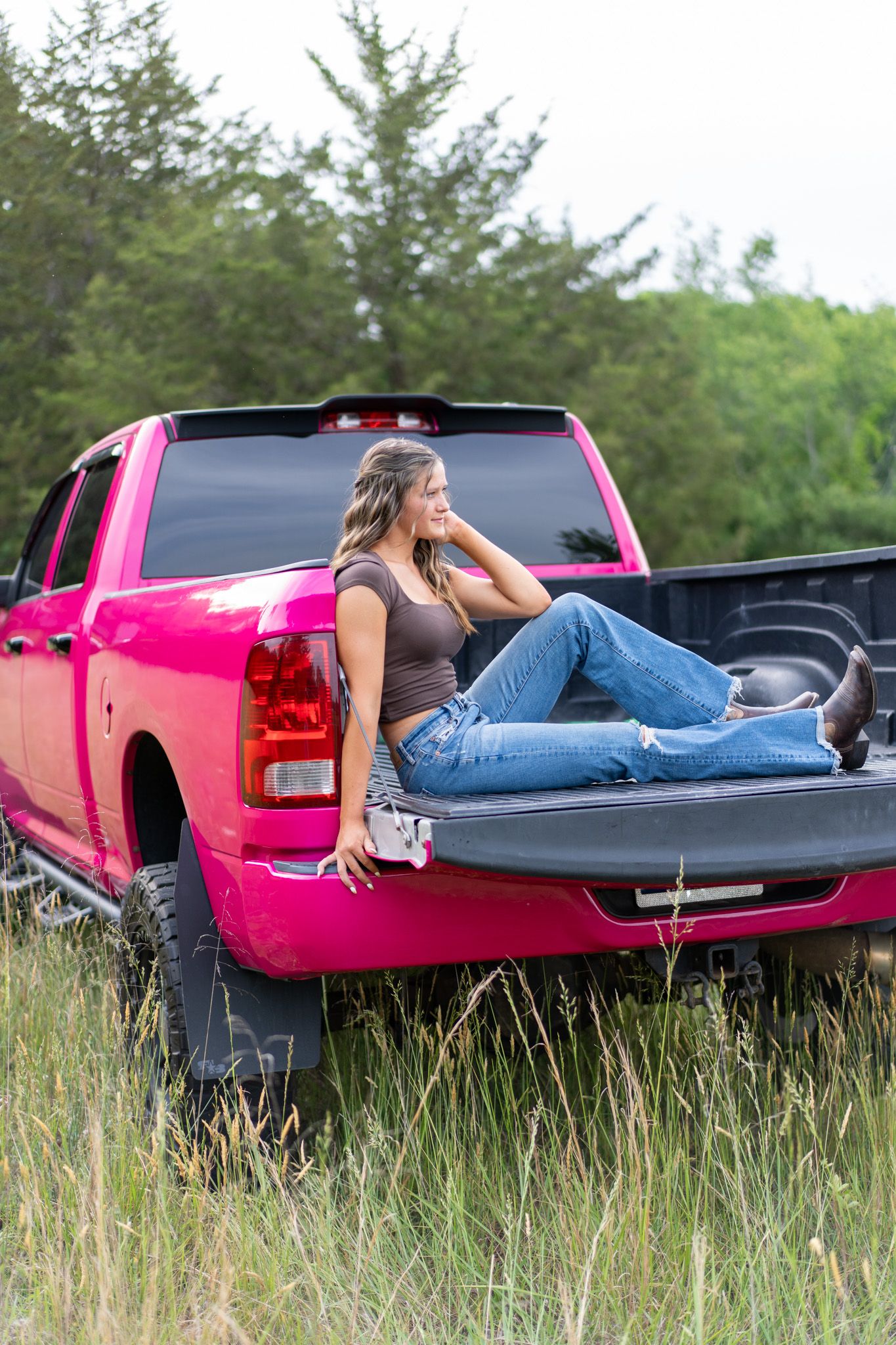 Young woman in jeans and boots relaxing in the bed of a bright pink pickup truck in a grassy field.