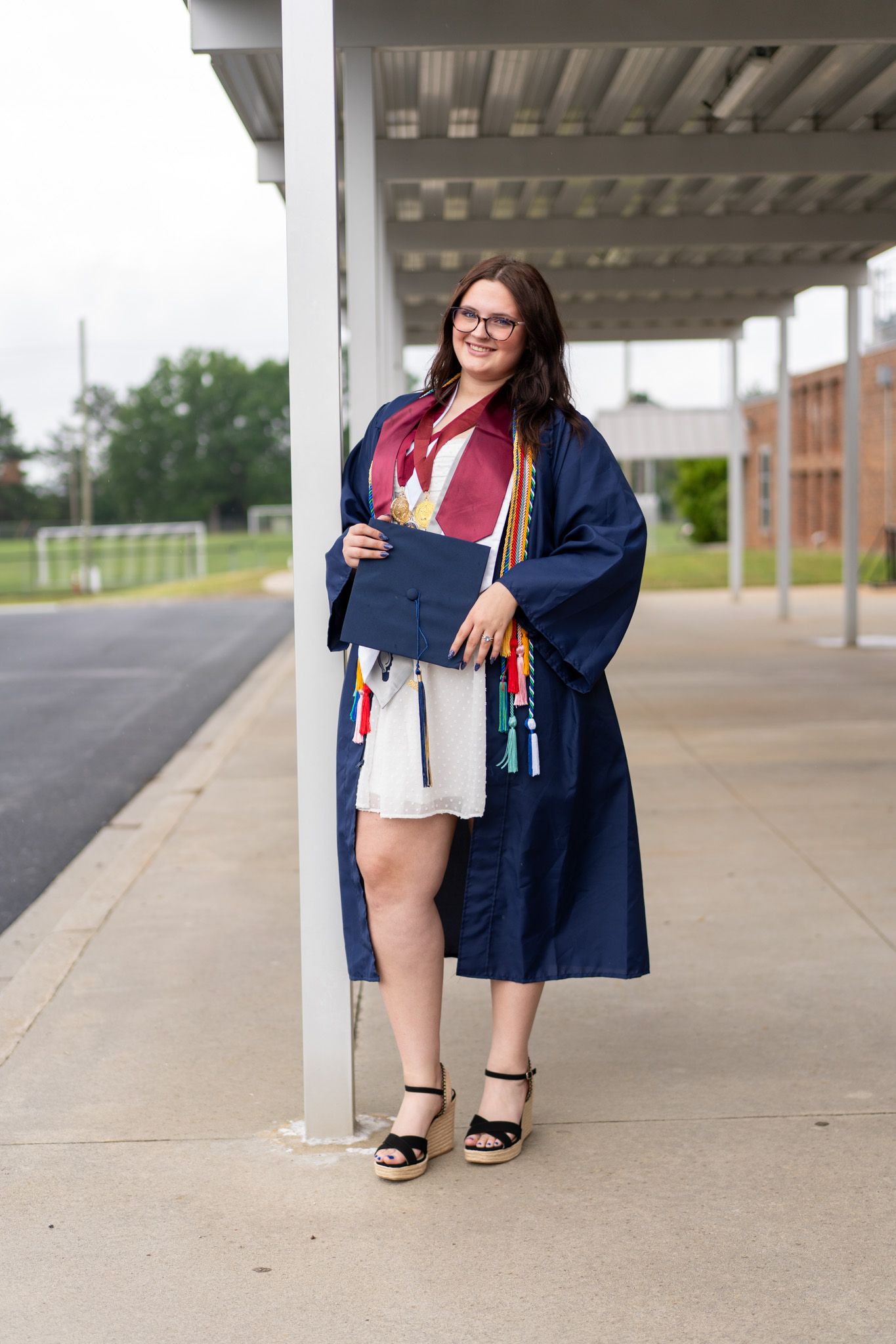 A smiling graduate in navy cap and gown holds diploma outdoors, wearing glasses and wedge heels, adorned with colorful honor cords.