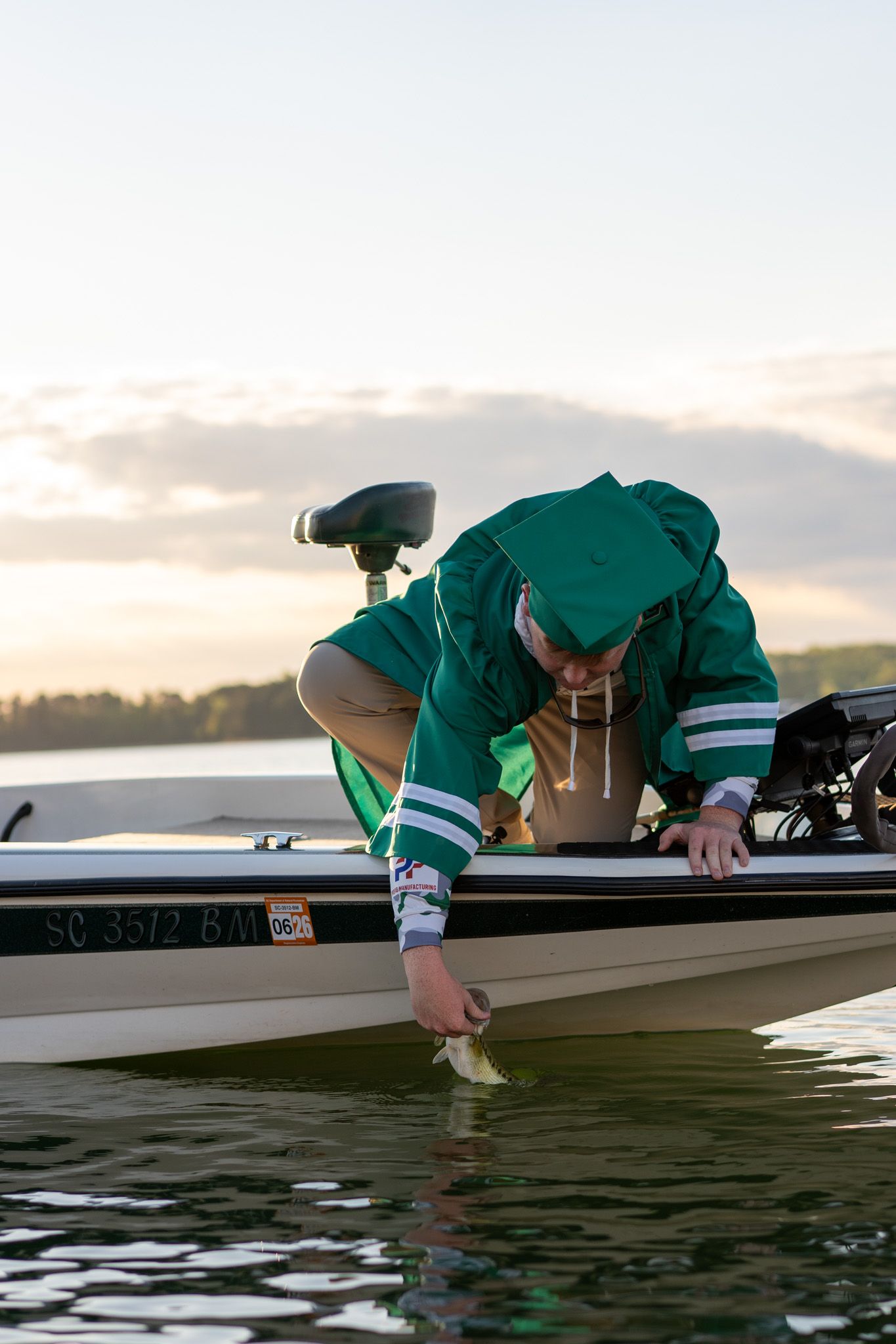 Angler in green jacket releasing a fish from a bass boat at sunset on a calm lake.