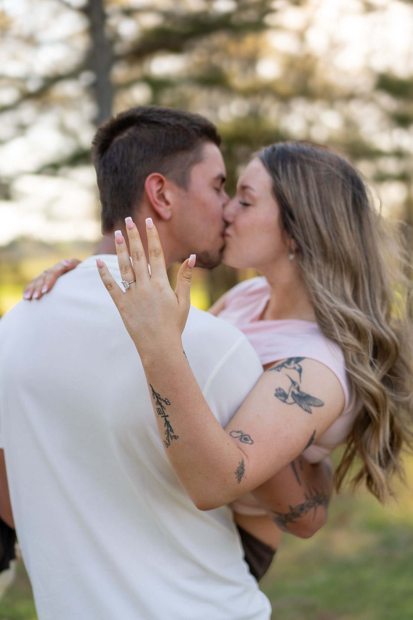 Engaged couple sharing a kiss outdoors with ring and tattoos visible in natural light.