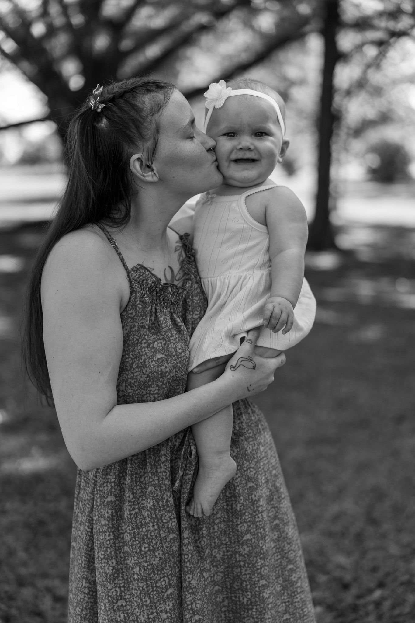 Black and white photo of young mother kissing laughing baby girl in white dress outdoors in park.