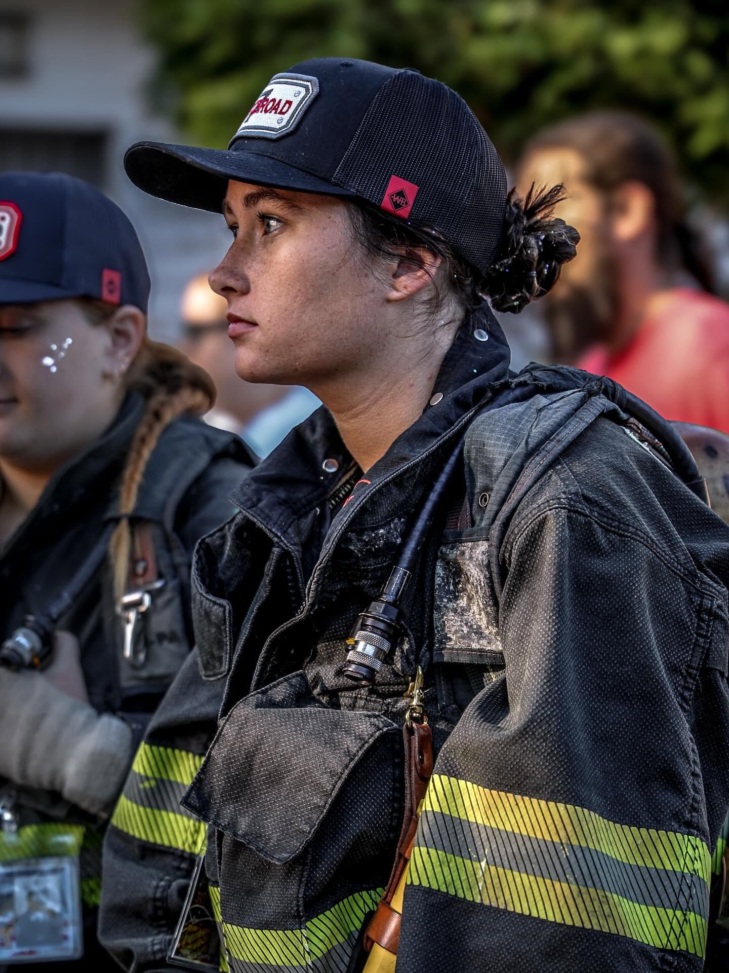 Female firefighter in full turnout gear and black cap standing attentively during outdoor fire training event.