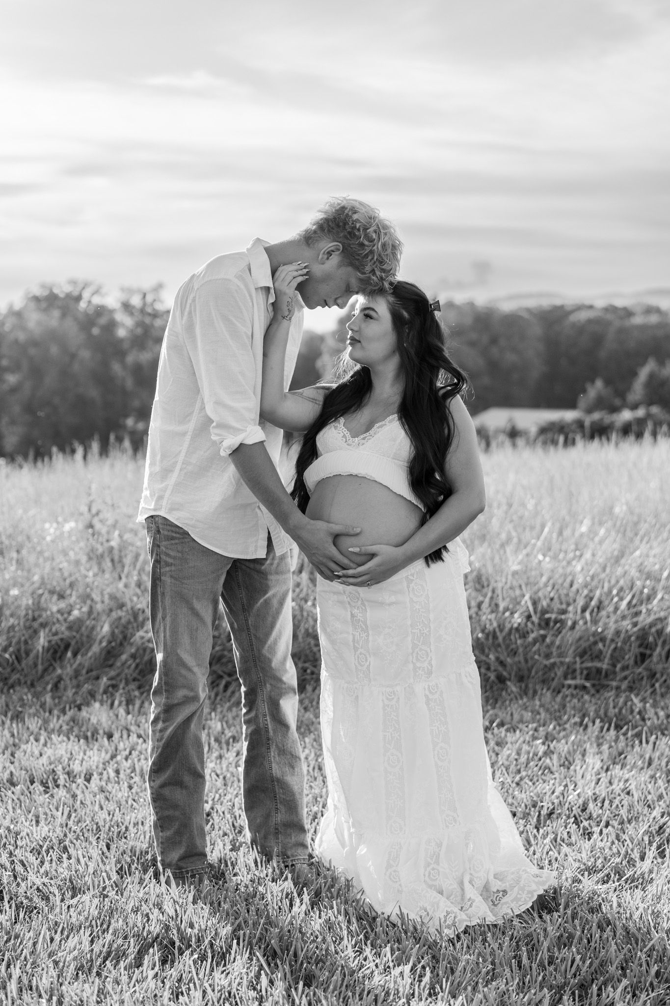 Black and white photo of couple tenderly embracing pregnant woman's baby bump in golden field.