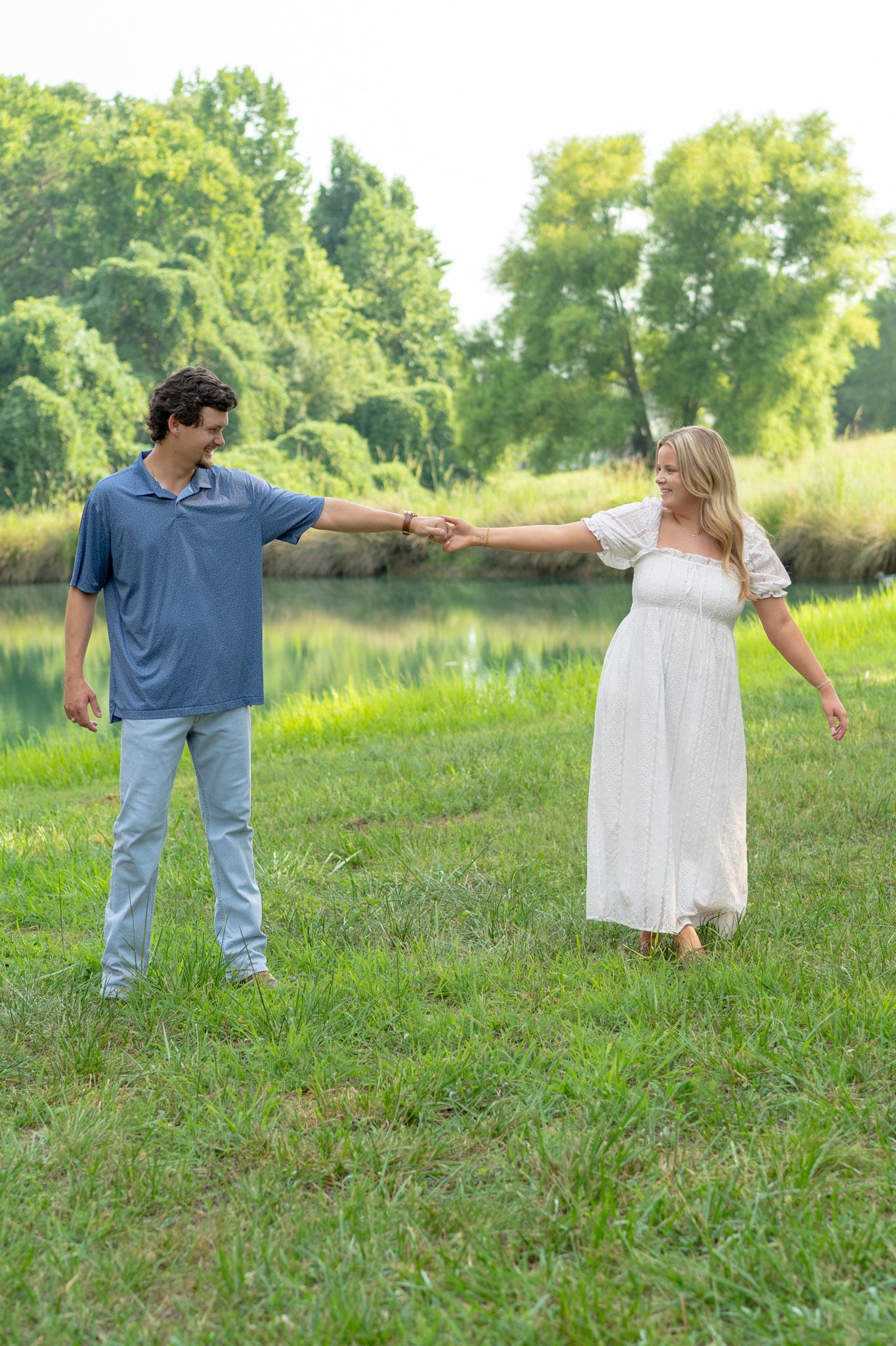 Couple holding hands and dancing on green grass near a pond surrounded by lush trees.