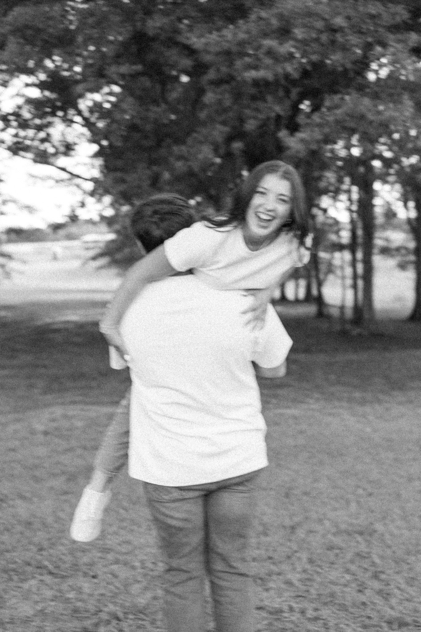 Black and white candid photo of laughing woman being carried piggyback by partner in outdoor park.