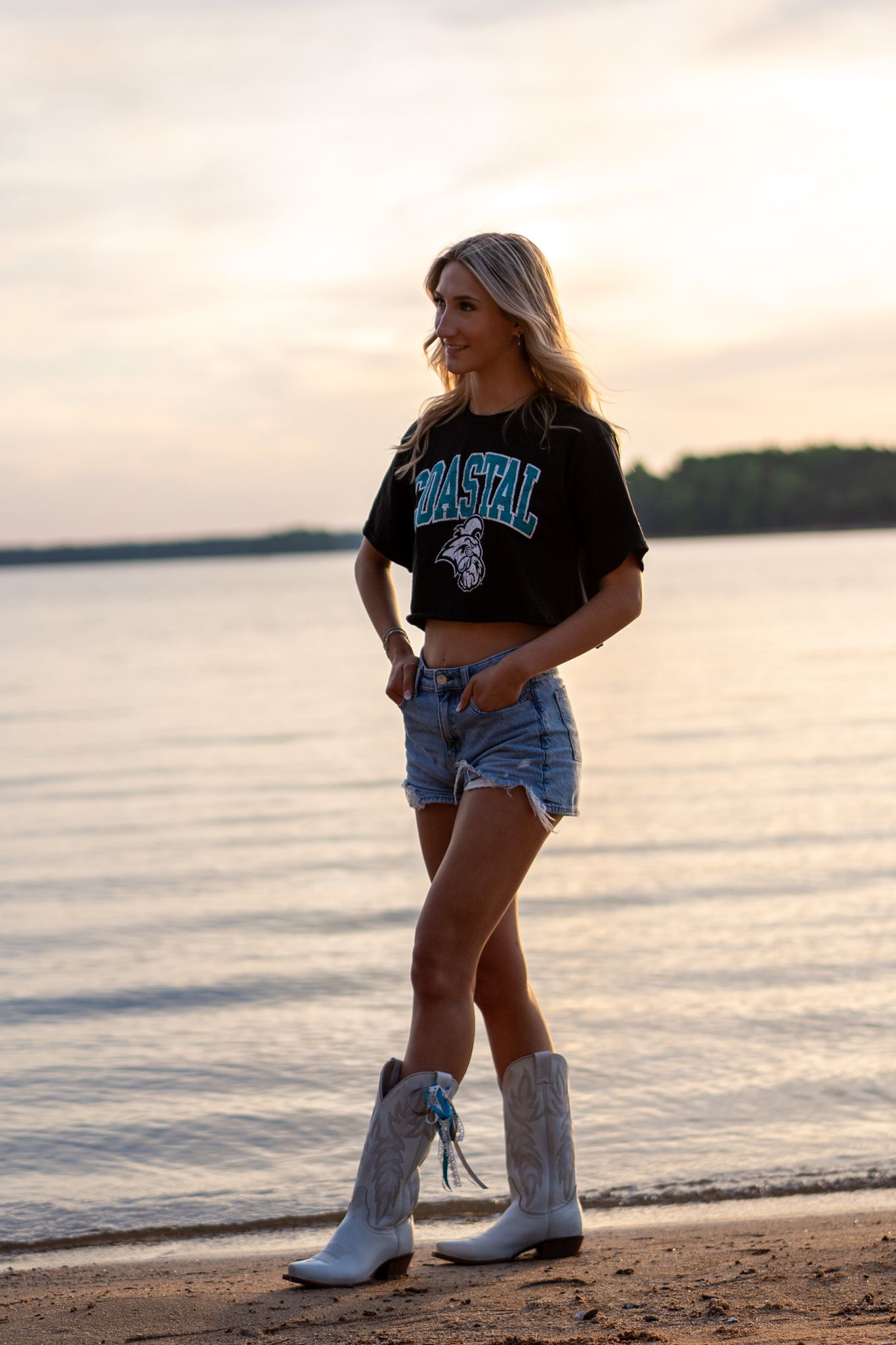 Blonde woman in black crop top, denim shorts, and white cowboy boots posing on a beach at sunset.