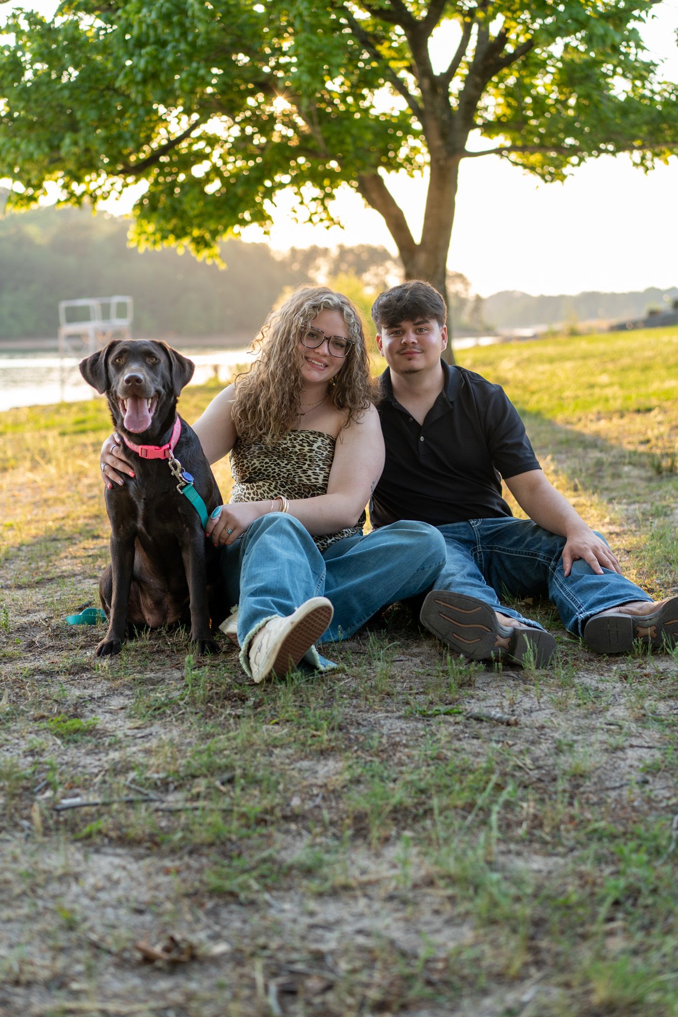Couple sitting on grass with chocolate Labrador at sunset near lake.