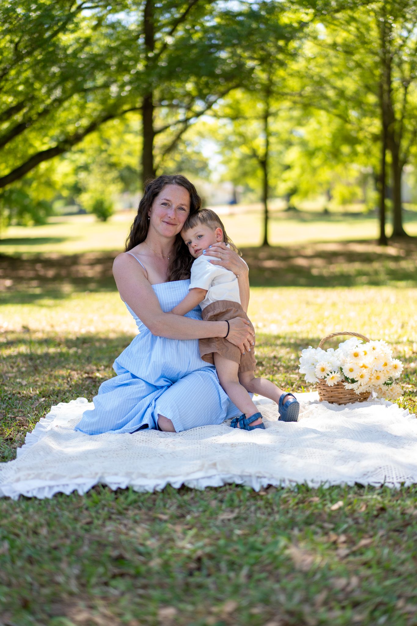 Mother in light blue dress cuddles toddler son on white blanket in sunny green park with flower basket.