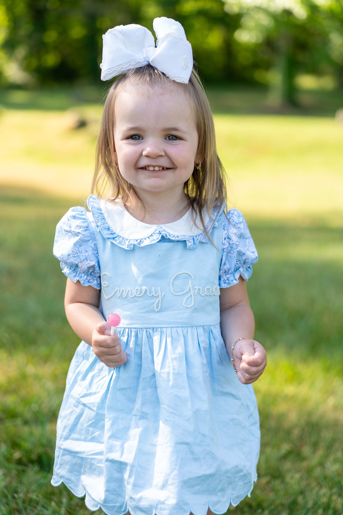 Adorable toddler girl in blue dress with white bow smiling in a sunny outdoor setting.