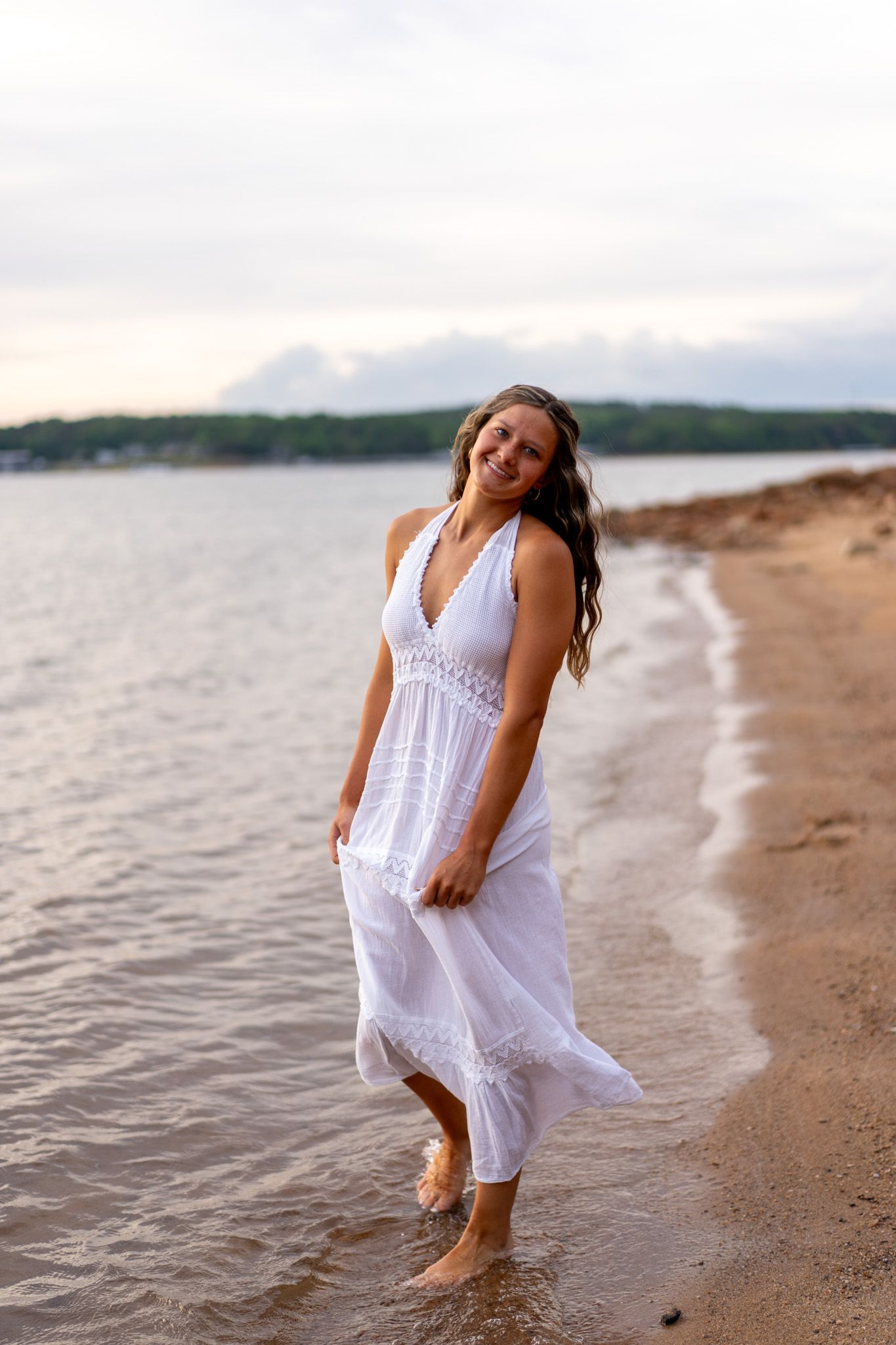 Smiling woman in white halter maxi dress walking barefoot on sandy beach shore at sunset.