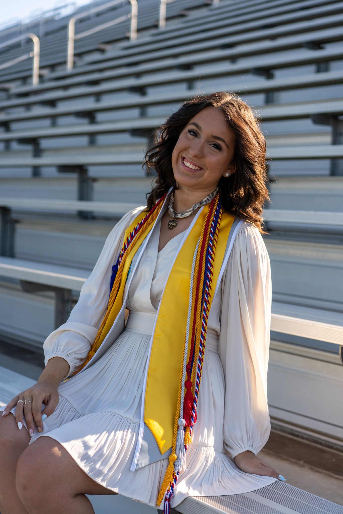 Smiling female graduate in white dress with gold honor stole sits in stadium bleachers for senior portrait.