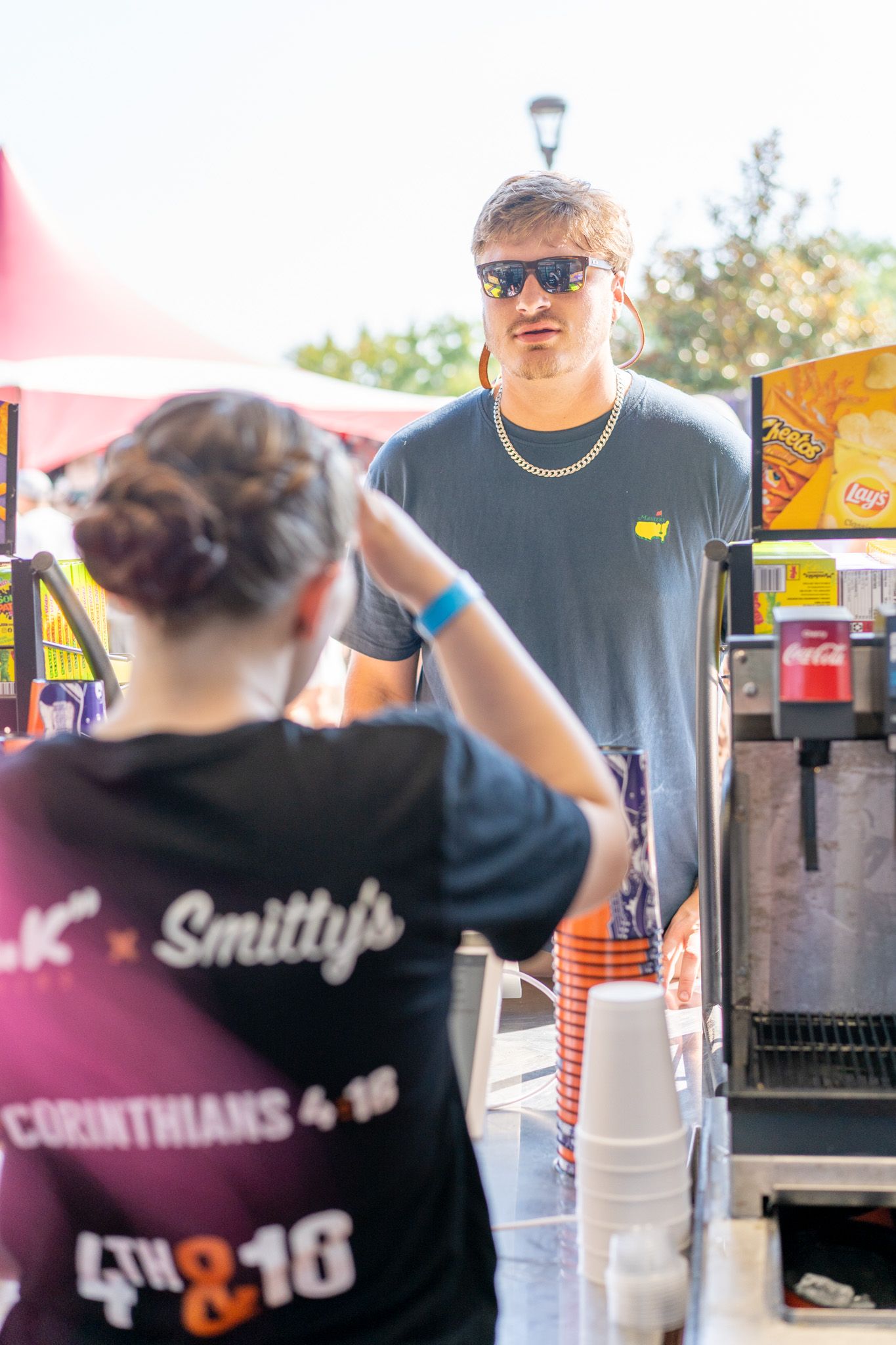 Man in Masters golf shirt and gold chain orders food at Smitty's outdoor festival concession stand on sunny day.