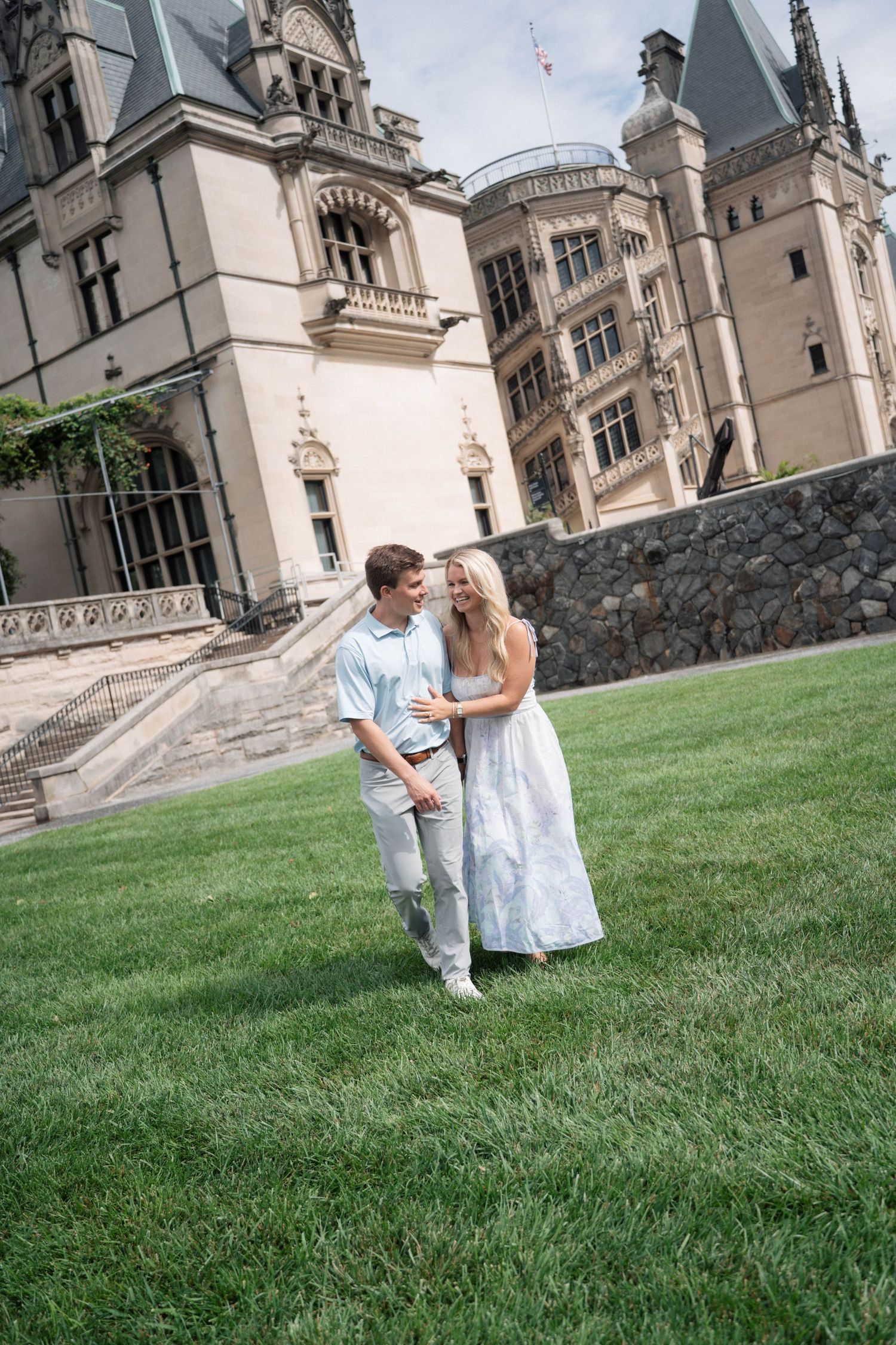 Smiling couple walking hand-in-hand on green lawn in front of grand historic estate building.