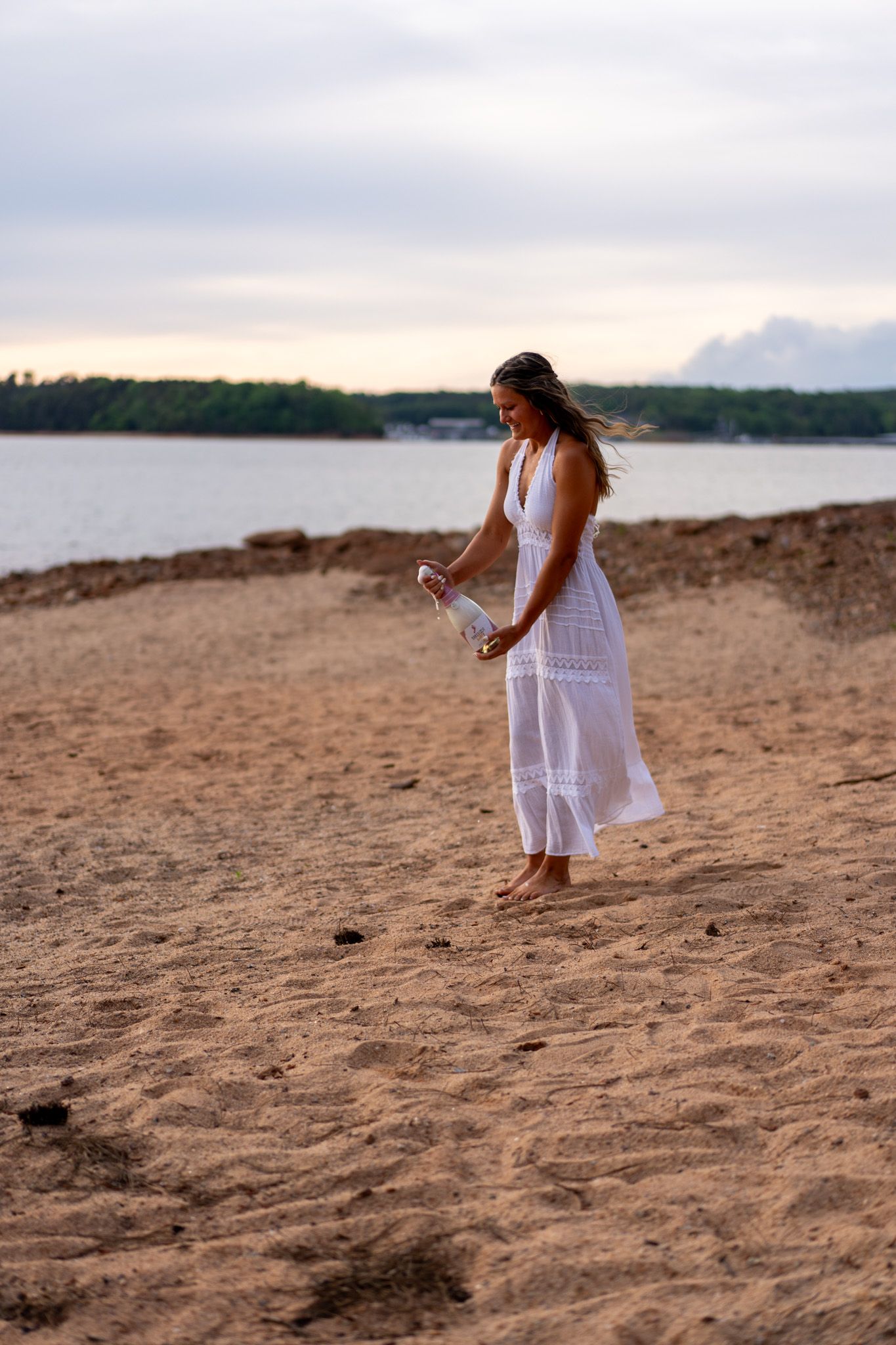 A woman in a white maxi dress standing barefoot on a sandy beach holding a champagne bottle at dusk.
