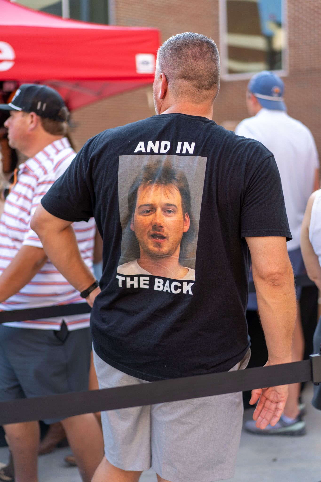 Man wearing black t-shirt with mugshot photo print reading 'And In The Back' at outdoor event.