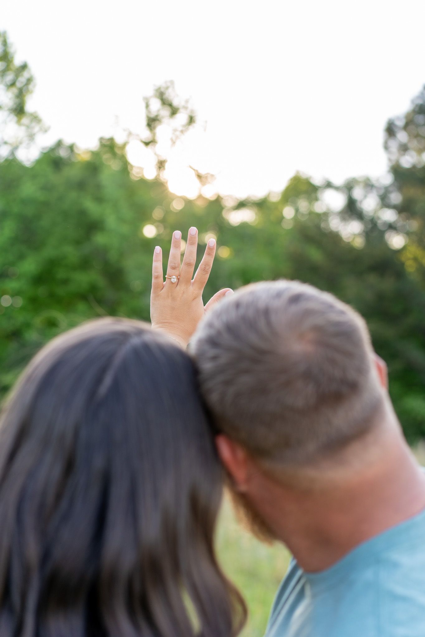 Couple showing off engagement ring outdoors with sunlit trees in background.