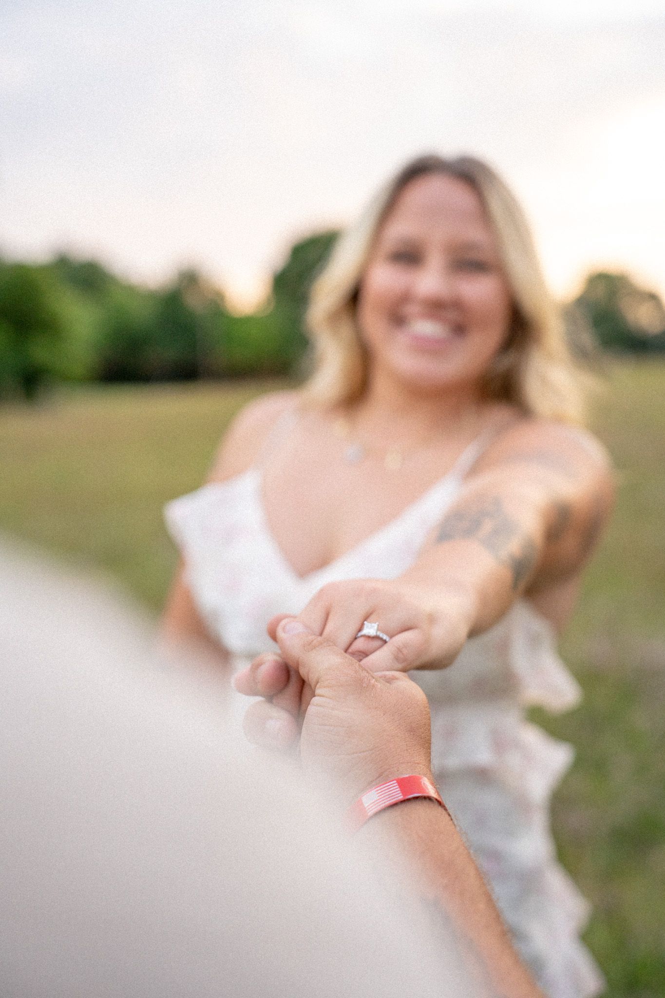 Smiling tattooed blonde woman in white dress extending hand with engagement ring toward camera at sunset.