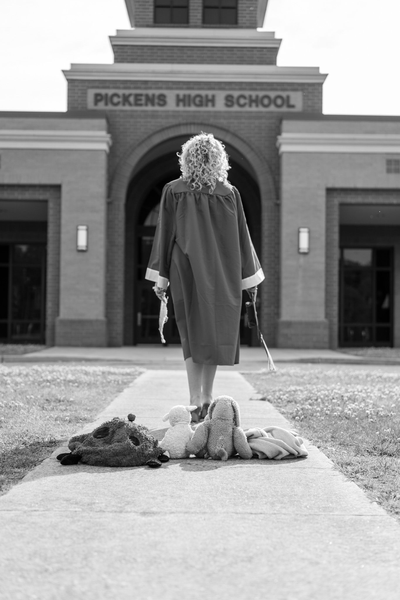 Graduate in cap and gown standing before Pickens High School entrance in black and white.