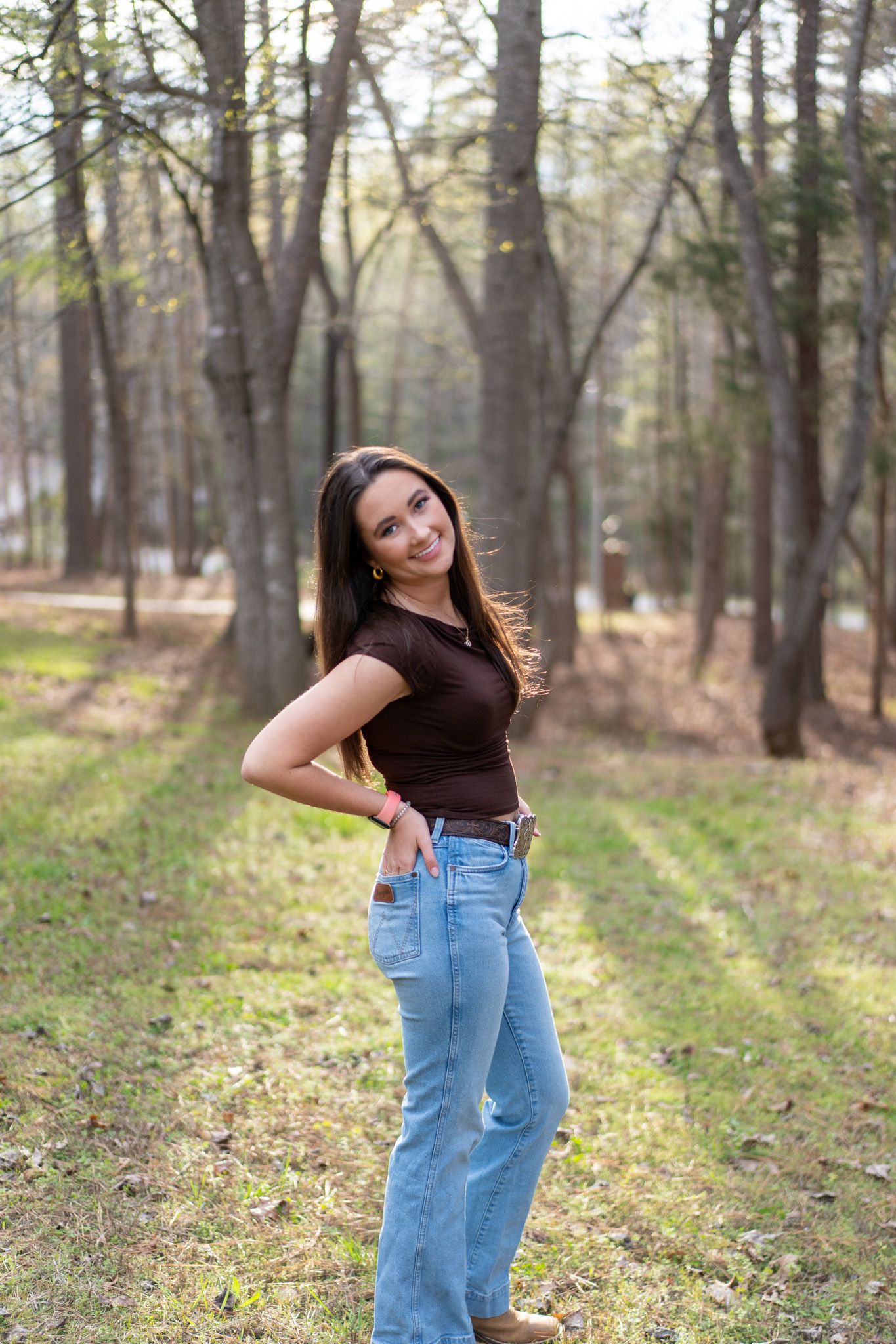 Smiling young woman in brown top and jeans posing in sunlit forest path.